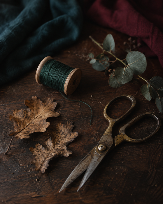 Herbstliches Stillleben mit Stoffschere, dunkelgrünem Nähgarn, Eichenlaub und Eukalyptus auf dunklem Holztisch